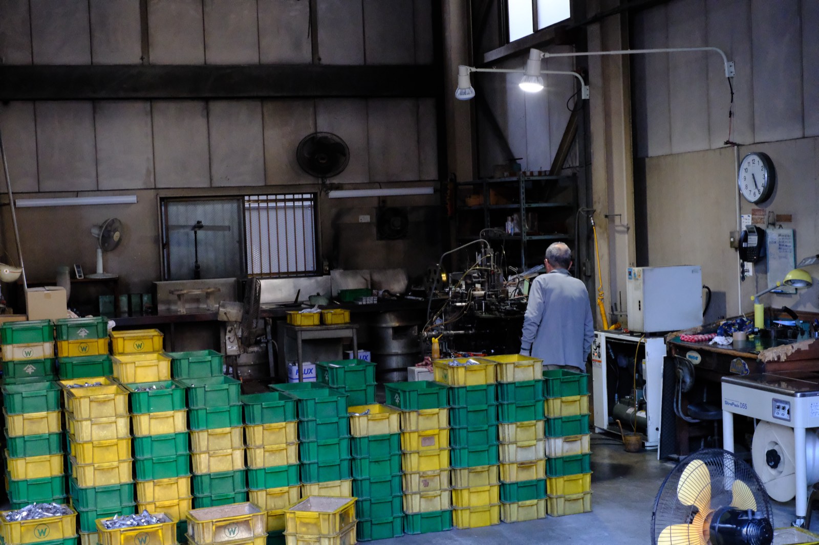 小さな町工場で一人、機械に向かって働く職人の後ろ姿。緑と黄色の部品ケースが積まれている。